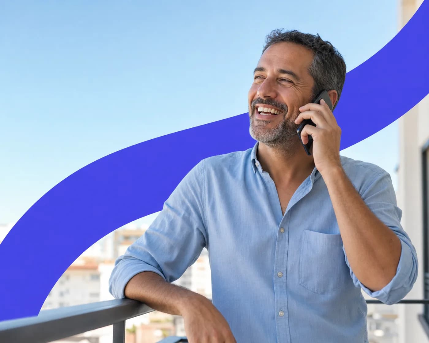 A man laughing on a phone call as he leans on a balcony railing under a blue sky