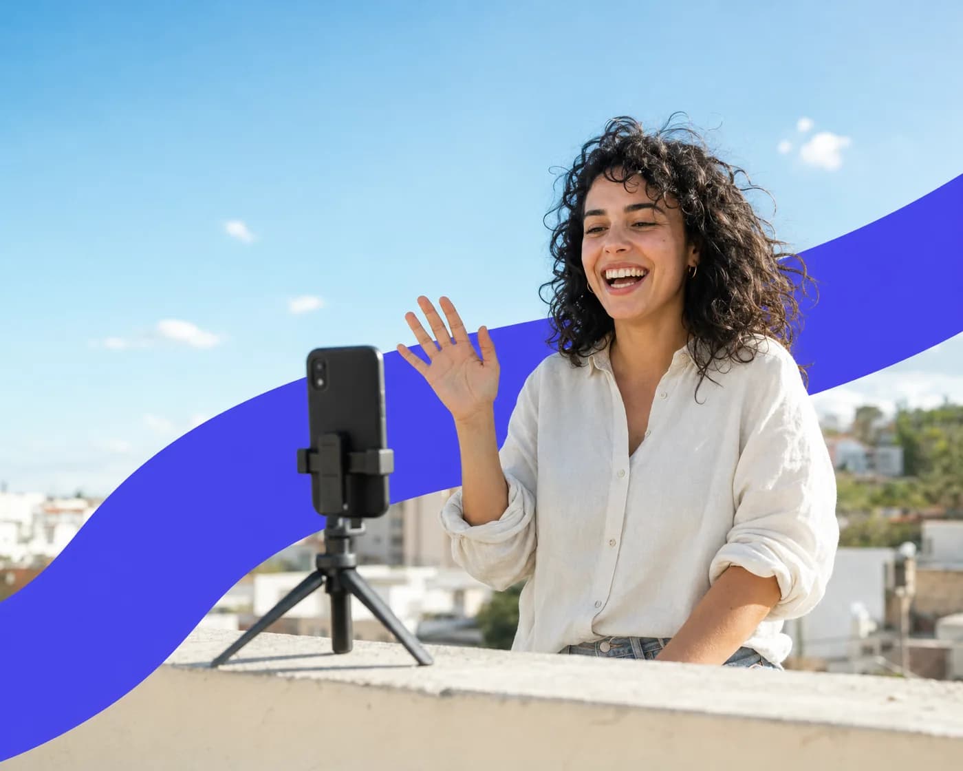 A smiling woman on a sunny rooftop waving as she films herself with a phone on a small tripod