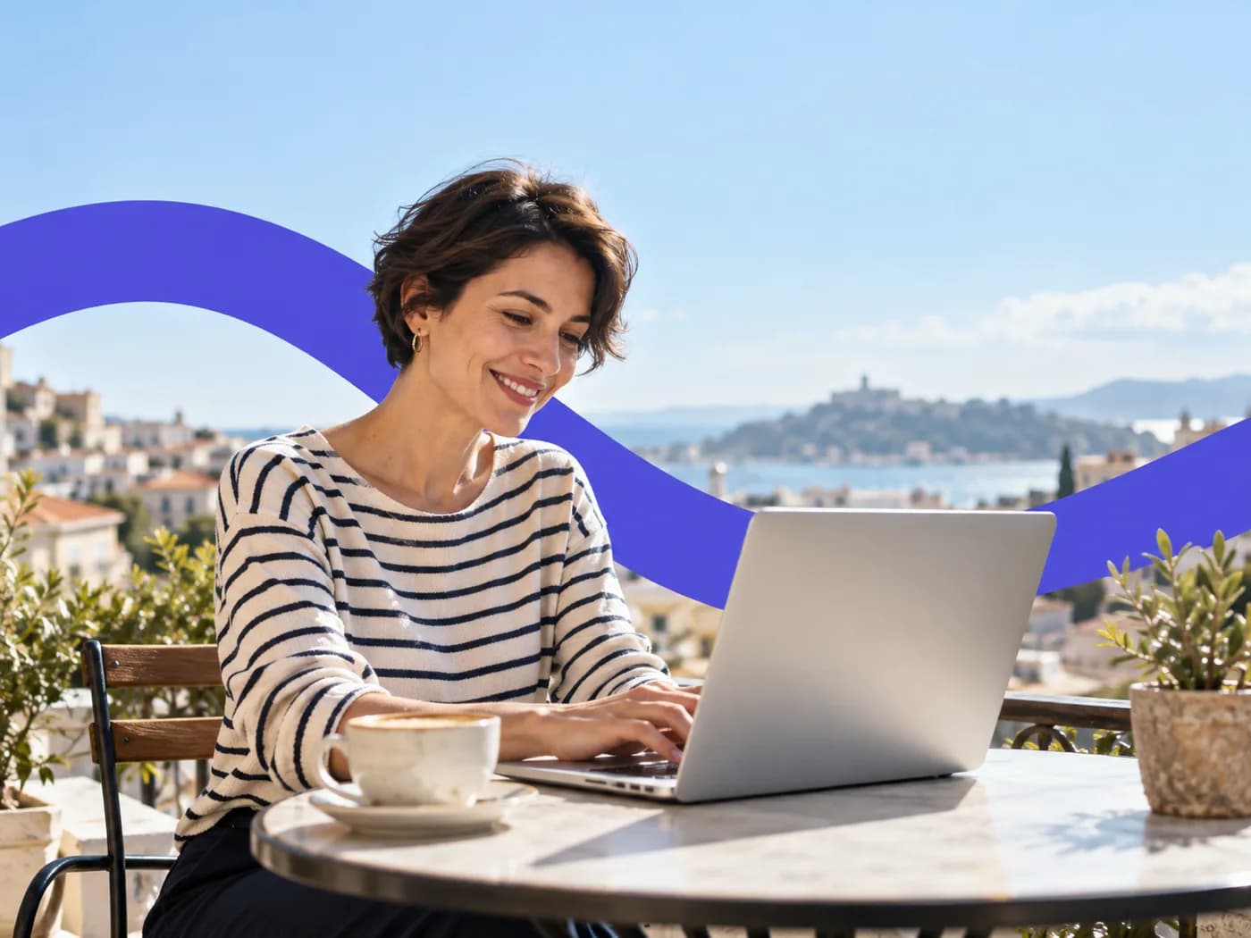 A woman smiling as she types on a laptop at a sunny cafe table with a coffee