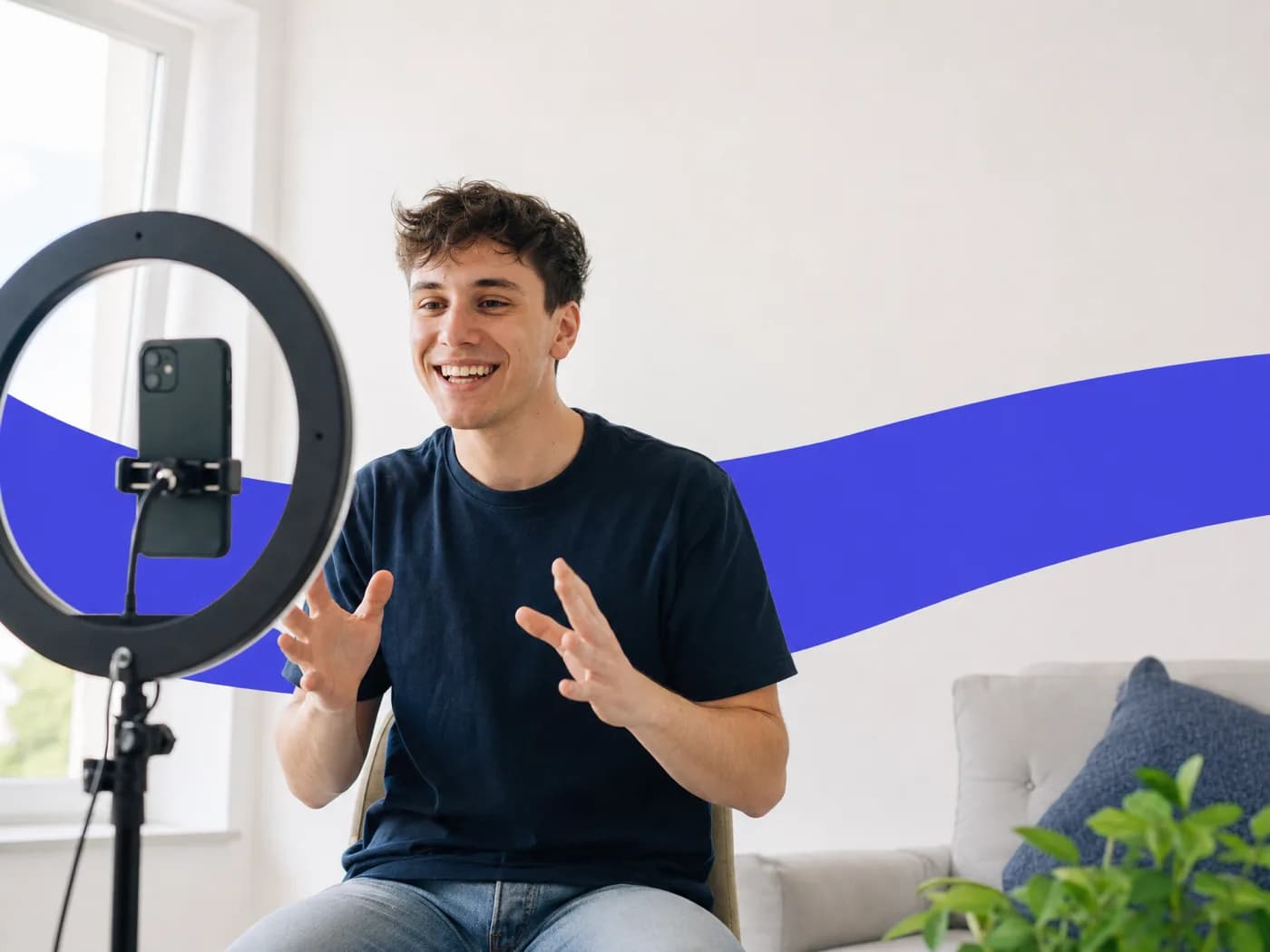 A smiling young man talking to a phone on a ring light stand in a bright room