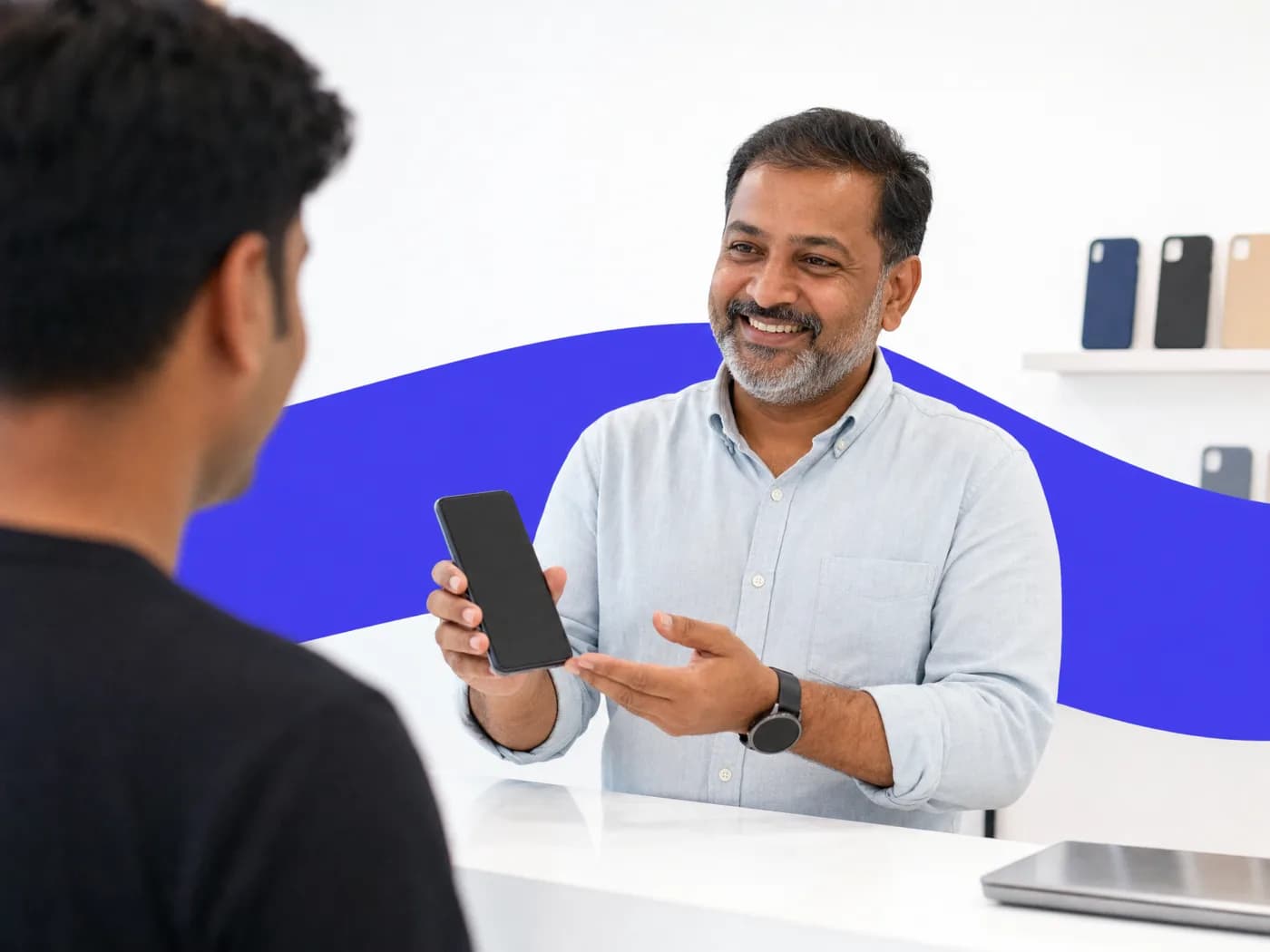 A phone shop owner smiling as he shows a phone to a customer across a bright counter