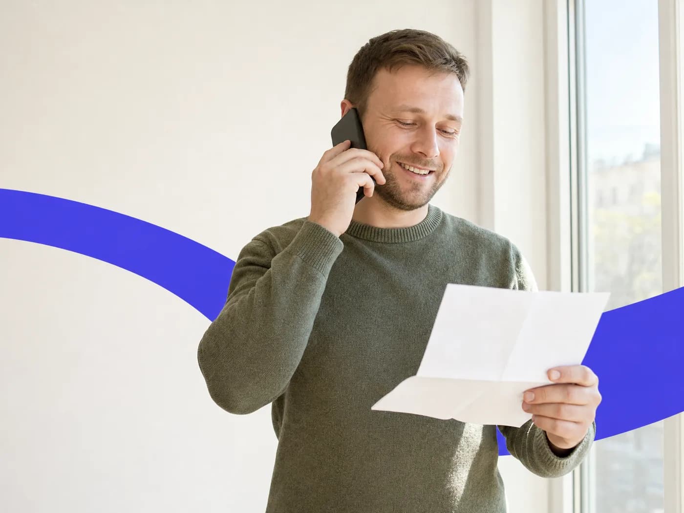 A man smiling with relief on a phone call by a sunny window, a folded letter in his other hand