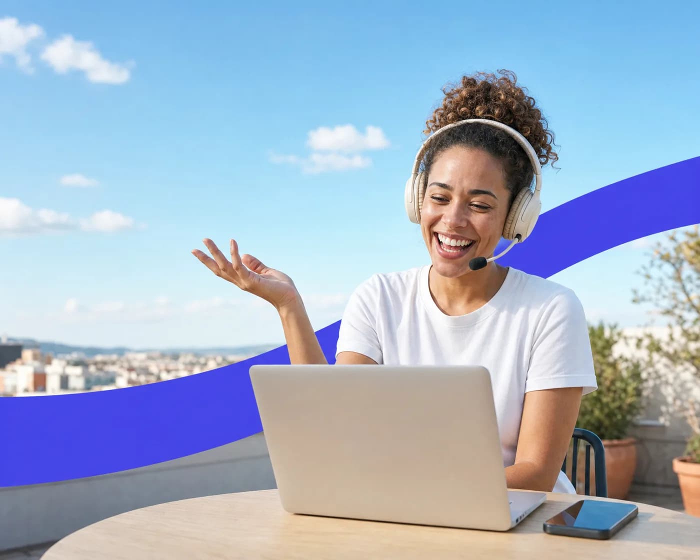 A woman laughing on a headset call at her laptop on a sunny rooftop, with her phone on the table beside it