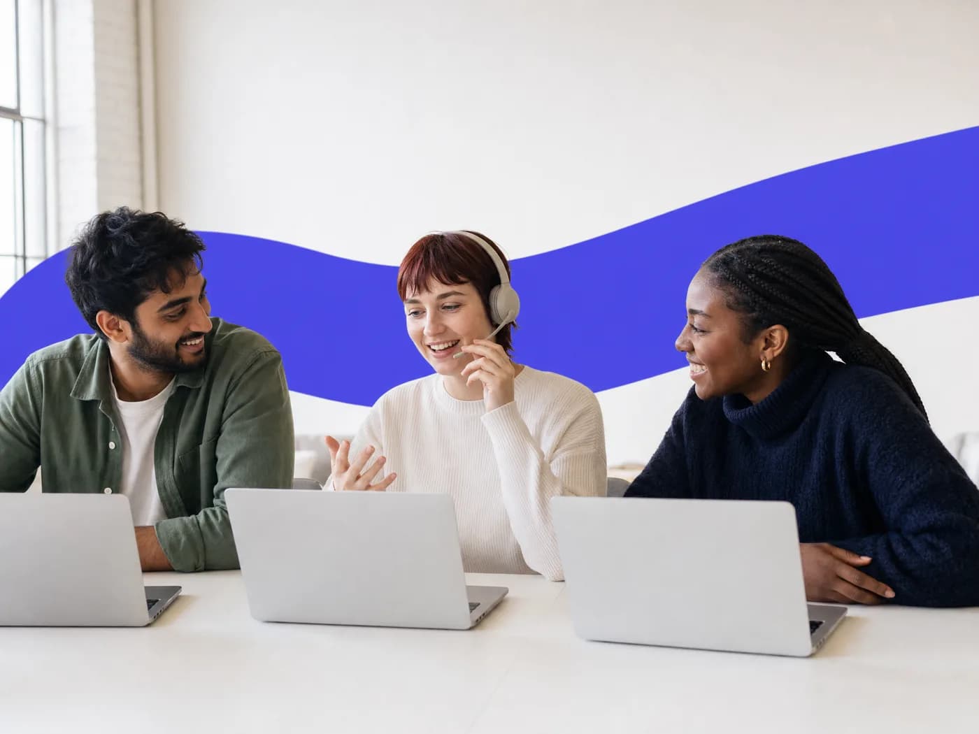 Three colleagues smiling at their laptops while one of them talks on a headset